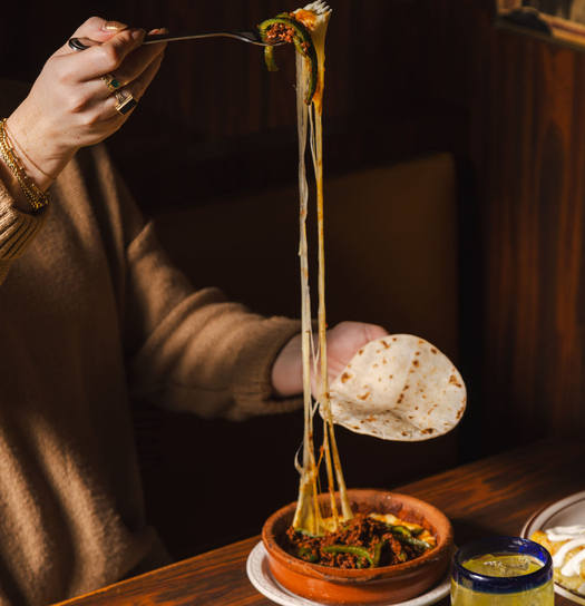 Person dipping tortilla chip into a bowl of chili topped with melted cheese and green peppers.