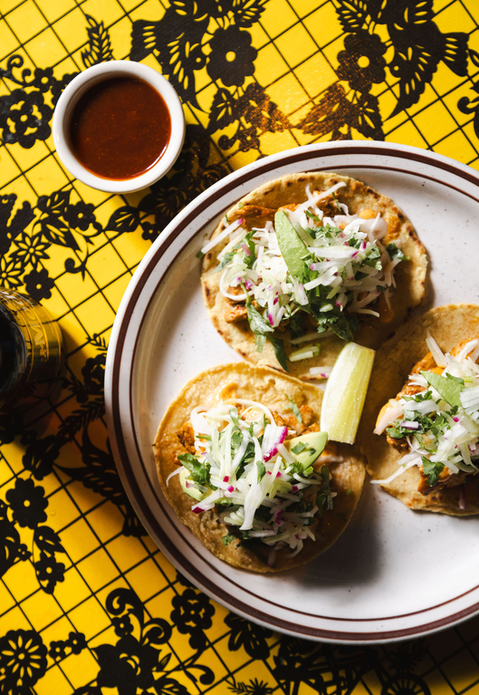Three crispy tacos on a plate with slaw, alongside a small dish of sauce and a glass of water on a yellow and black patterned tablecloth.