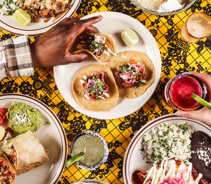 Colorful Mexican dishes on a vibrant tablecloth, includes tacos, guacamole, and a drink.
