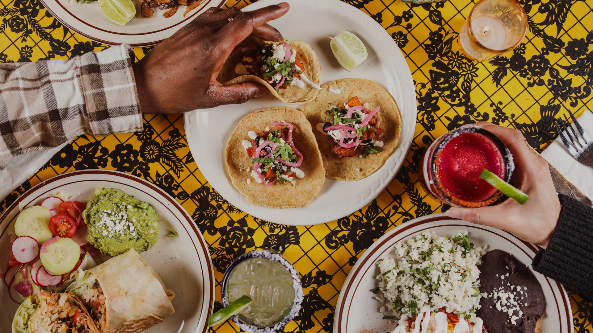 A vibrant restaurant setting with yellow and black patterned tablecloth, featuring tacos, a burrito, guacamole, rice, black beans, a margarita, and a lime wedge.