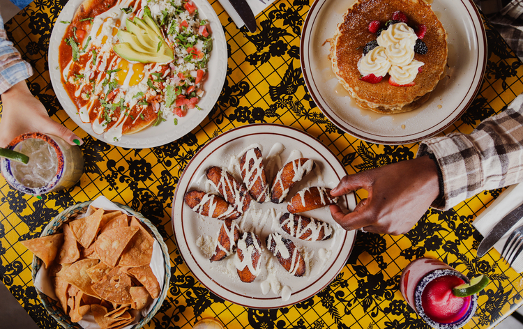 Diverse breakfast spread: huevos rancheros, pancakes, churros, chips, and drinks on a vibrant tablecloth.