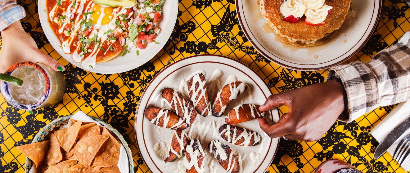Overhead view of a vibrant table with a yellow patterned tablecloth. Dishes include pancakes, nachos, and a creamy dessert with a drizzle. Hands are reaching for food.